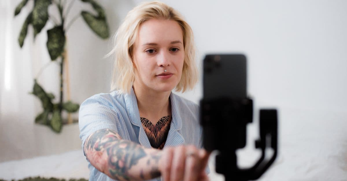 Tattooed woman with blonde hair adjusts smartphone on tripod for vlog setup indoors.