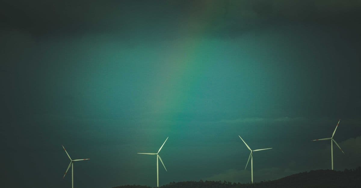 Wind turbines on a hill under a dramatic sky with visible rainbow, promoting renewable energy.