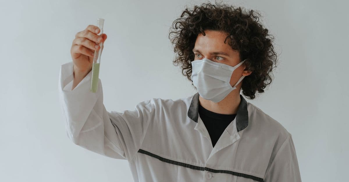 A scientist in a lab coat examining a test tube while wearing a face mask.