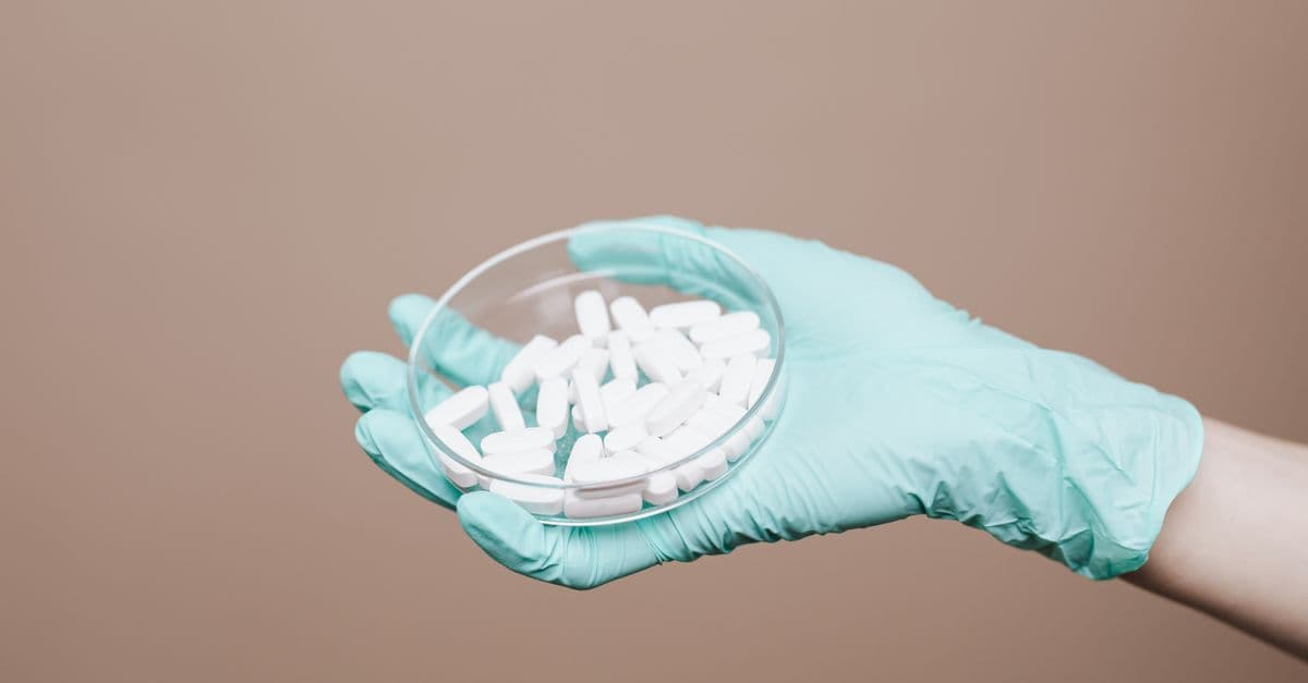 A gloved hand holding a petri dish filled with white pills in a healthcare setting.