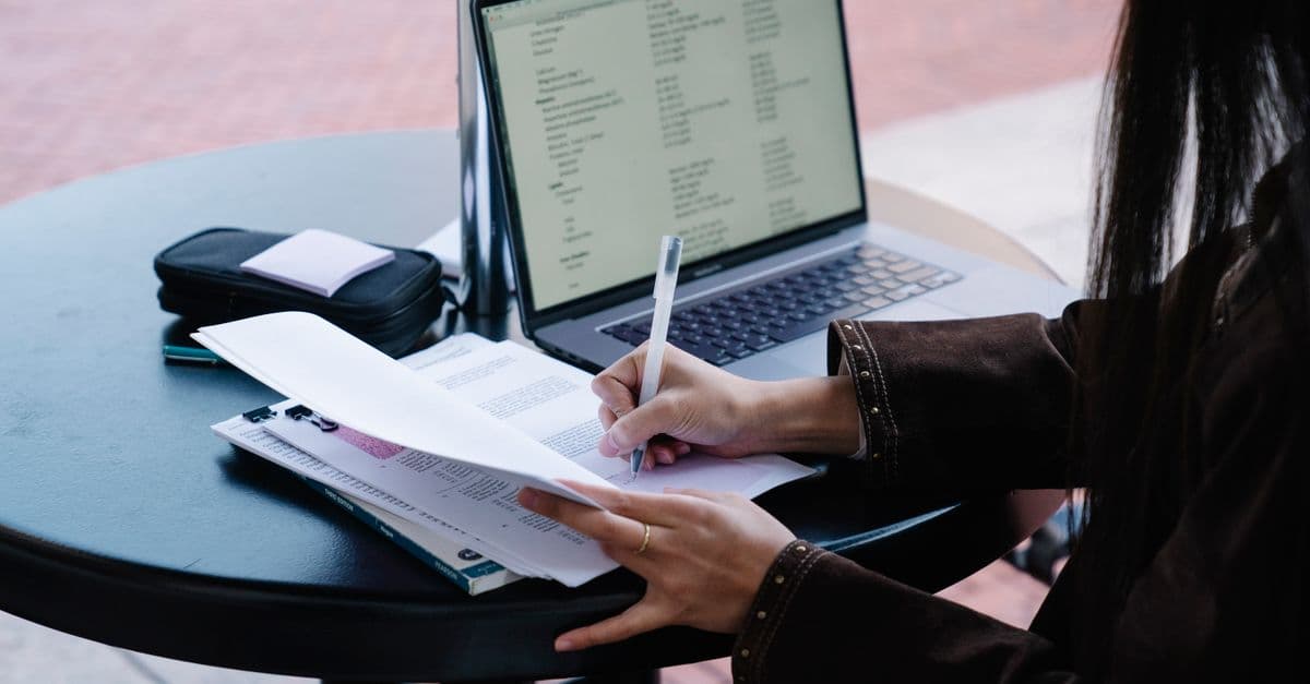 A woman writes on papers at an outdoor cafe table with an open laptop.