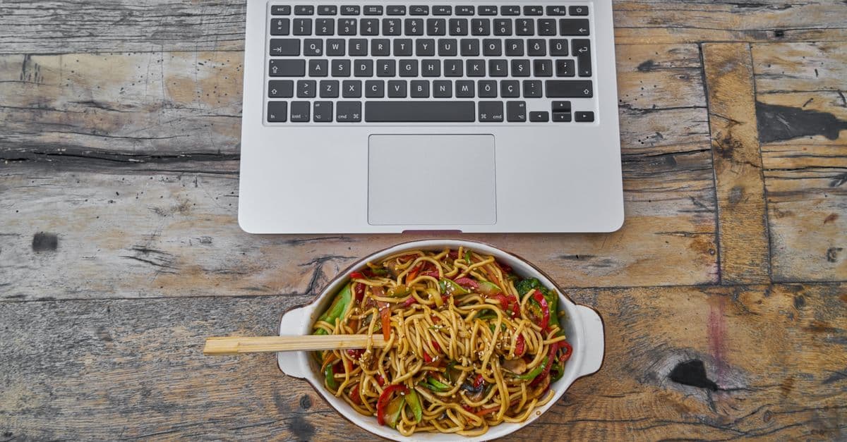 Overhead view of laptop and noodle bowl on rustic wooden table, perfect blend of food and technology.