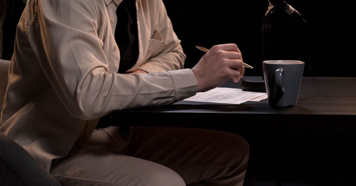 A man in a studio setting working on a podcast with microphone, pen, and coffee cup.