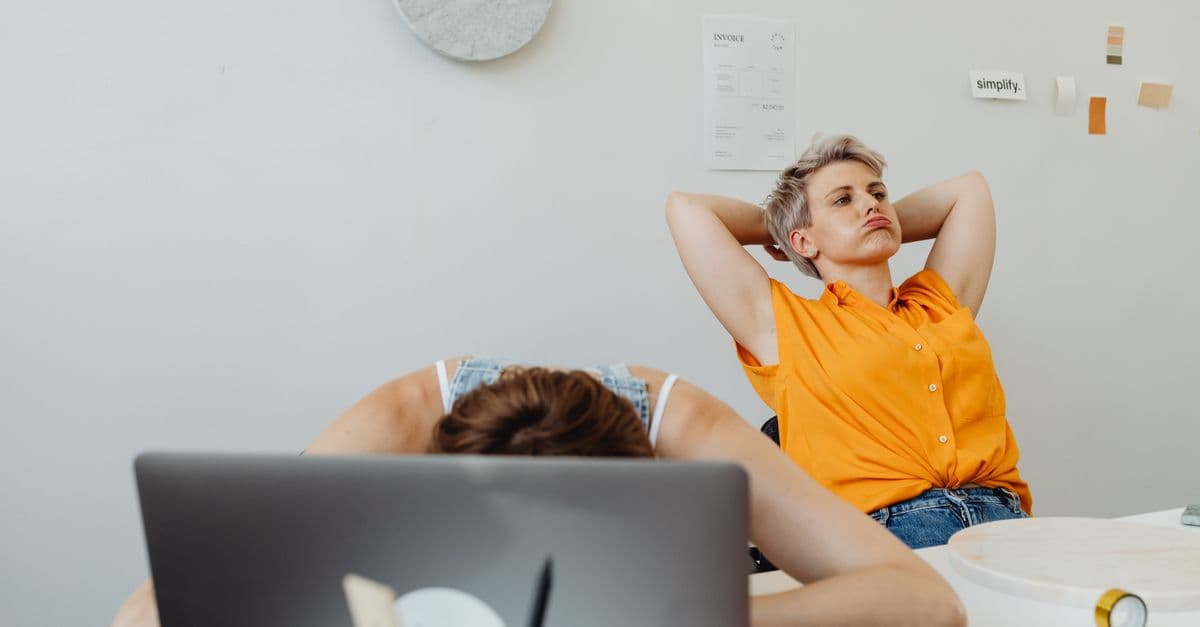 Two women in office, one stressed with head on desk, another relaxed. Modern workplace dynamics.