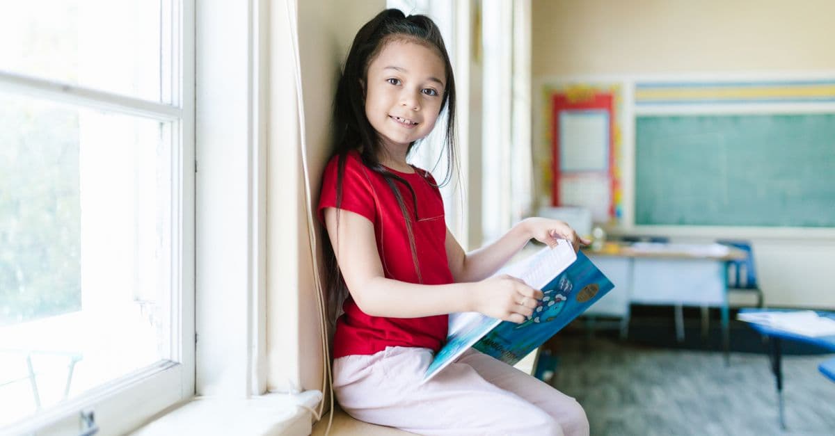 A young girl smiles while reading a book by the window in a bright classroom.