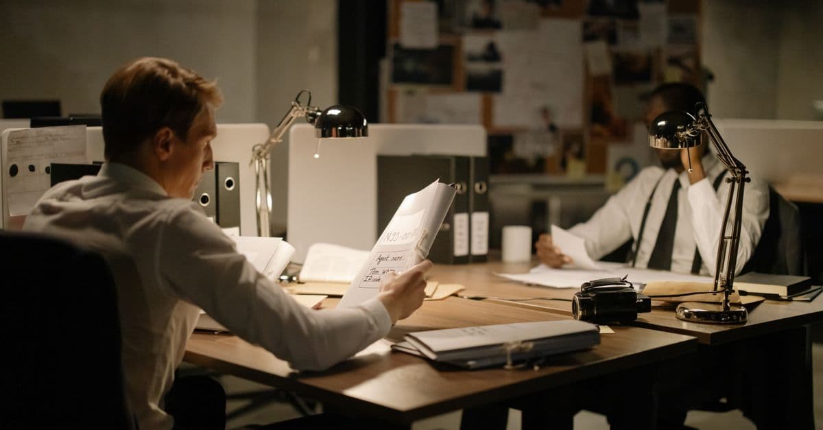 Two detectives analyzing documents in an office setting under dim lighting, focusing on investigation.