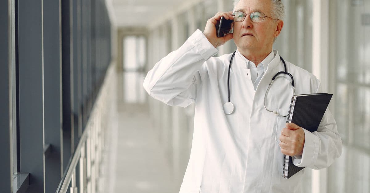 Serious mature male doctor in medical uniform standing with stethoscope and notebook while talking on smartphone in clinic corridor