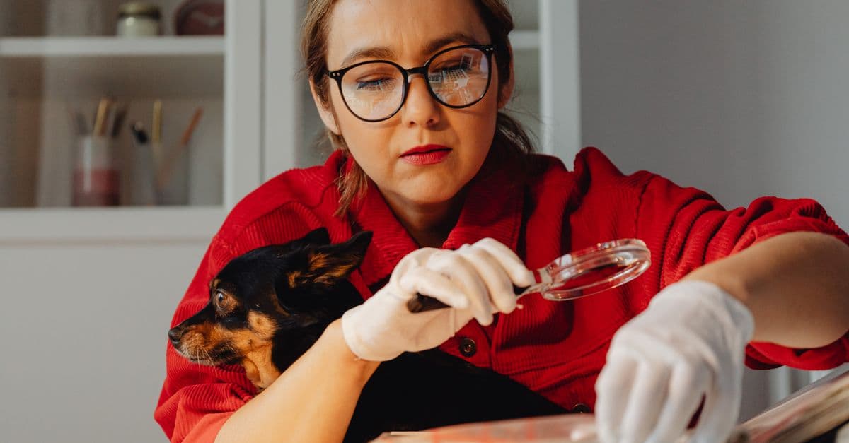 A woman wearing glasses examines documents with a magnifying glass, holding her dog.