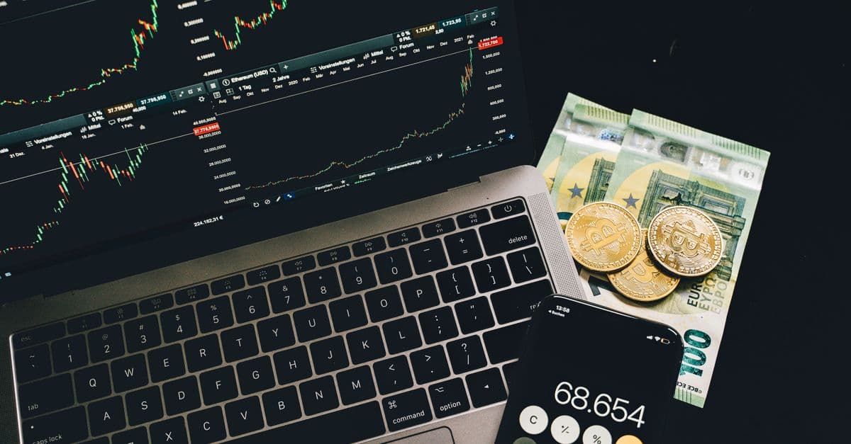 A laptop displaying stock charts with Bitcoin, Euros, and a cellphone calculator, showcasing financial analysis.