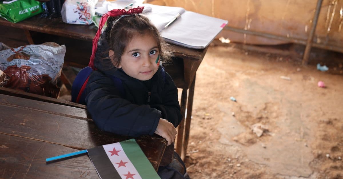A young girl in a Syrian classroom holding a flag and smiling.