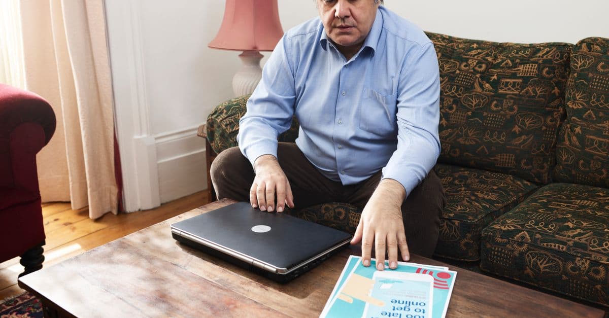A middle-aged man sitting on a couch using a laptop and looking at a flyer on a coffee table.