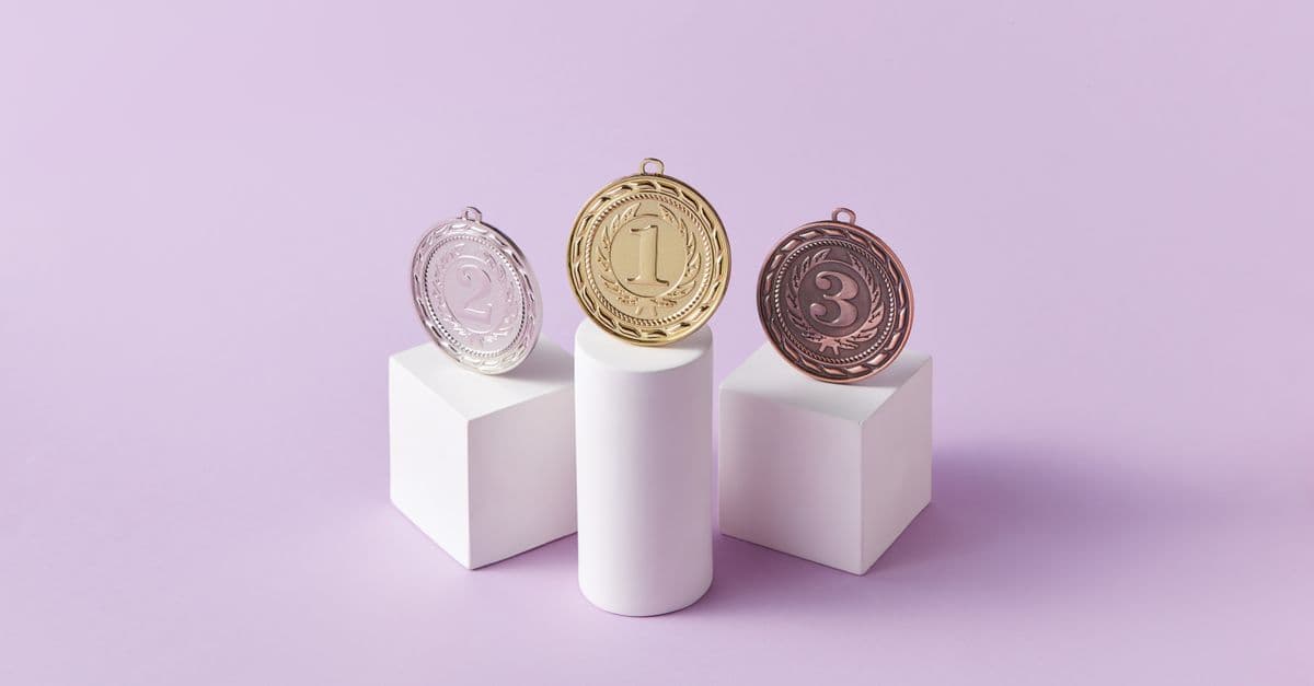 Gold, silver, and bronze medals displayed on white pedestals with a pink background.
