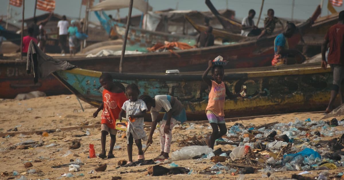 Children play amidst trash on a polluted beach with fishing boats in the background.
