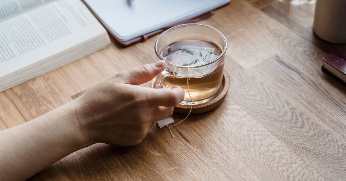 A serene setting with a hand holding a cup of tea, accompanied by a notepad and book on a wooden table.
