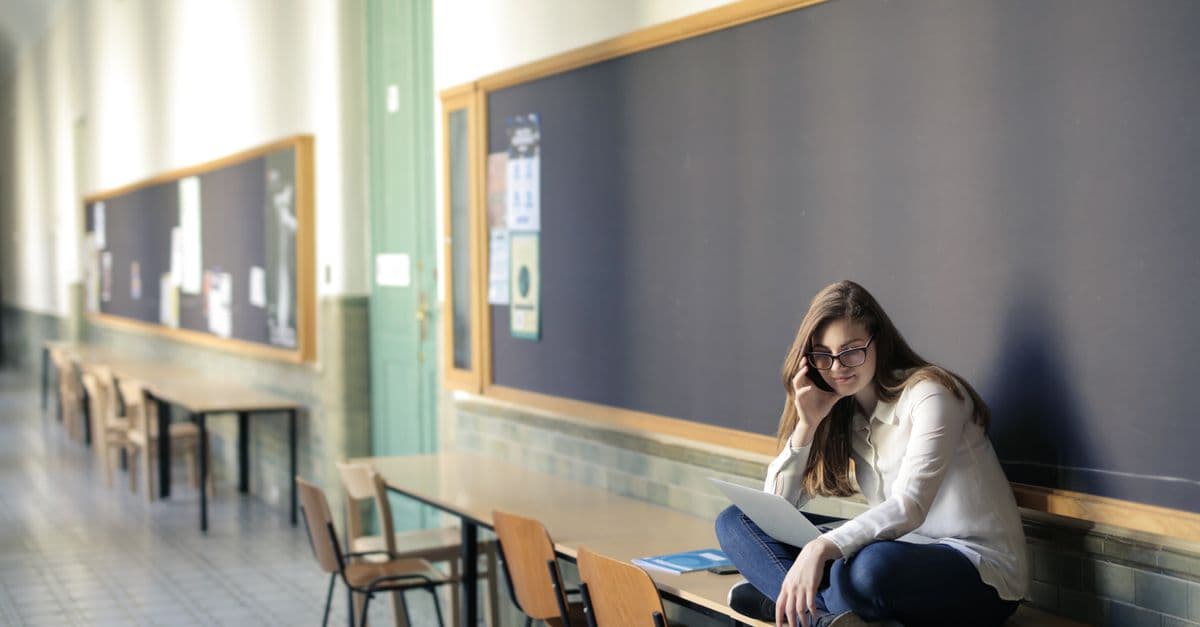 A woman sits on a desk in a university hallway, studying on a laptop.