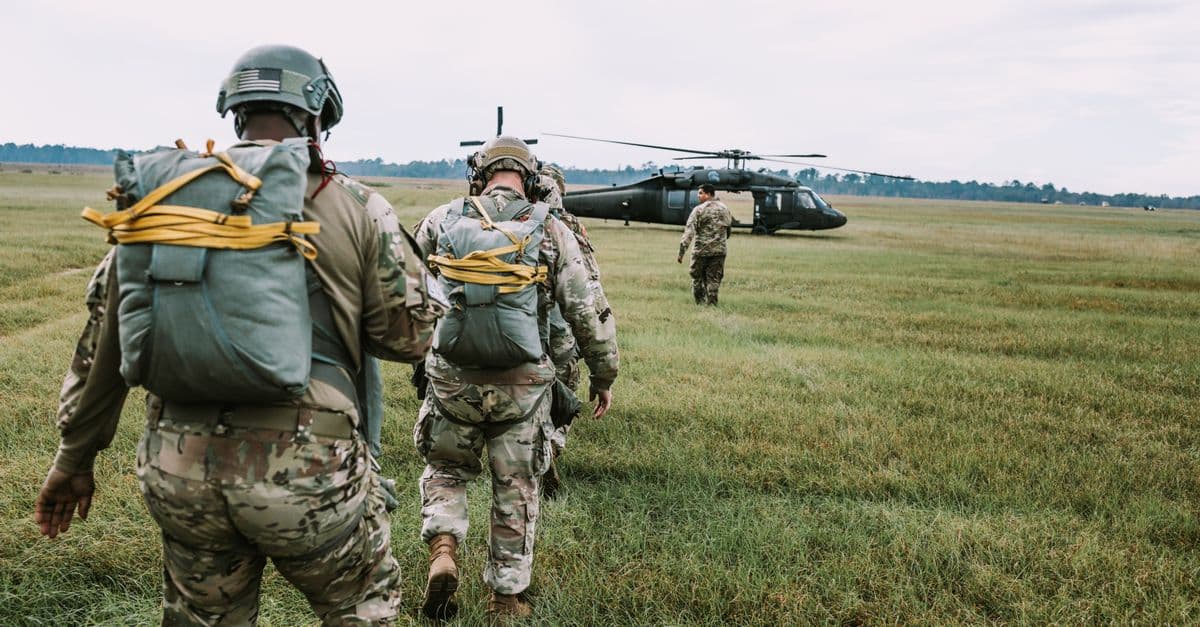 Military personnel in uniforms and gear walk toward a helicopter on a grassy field.
