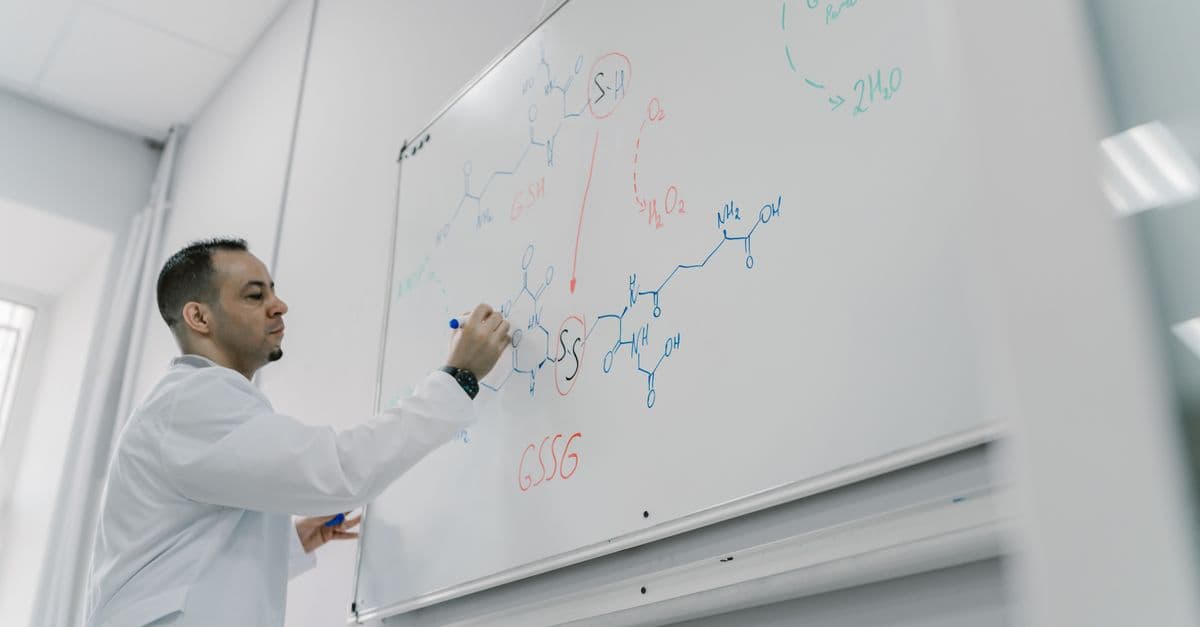 Male teacher explains chemical equation on whiteboard in science classroom setting.