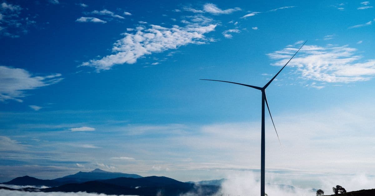 A solitary wind turbine rises against a vibrant blue sky, surrounded by clouds and distant mountains.
