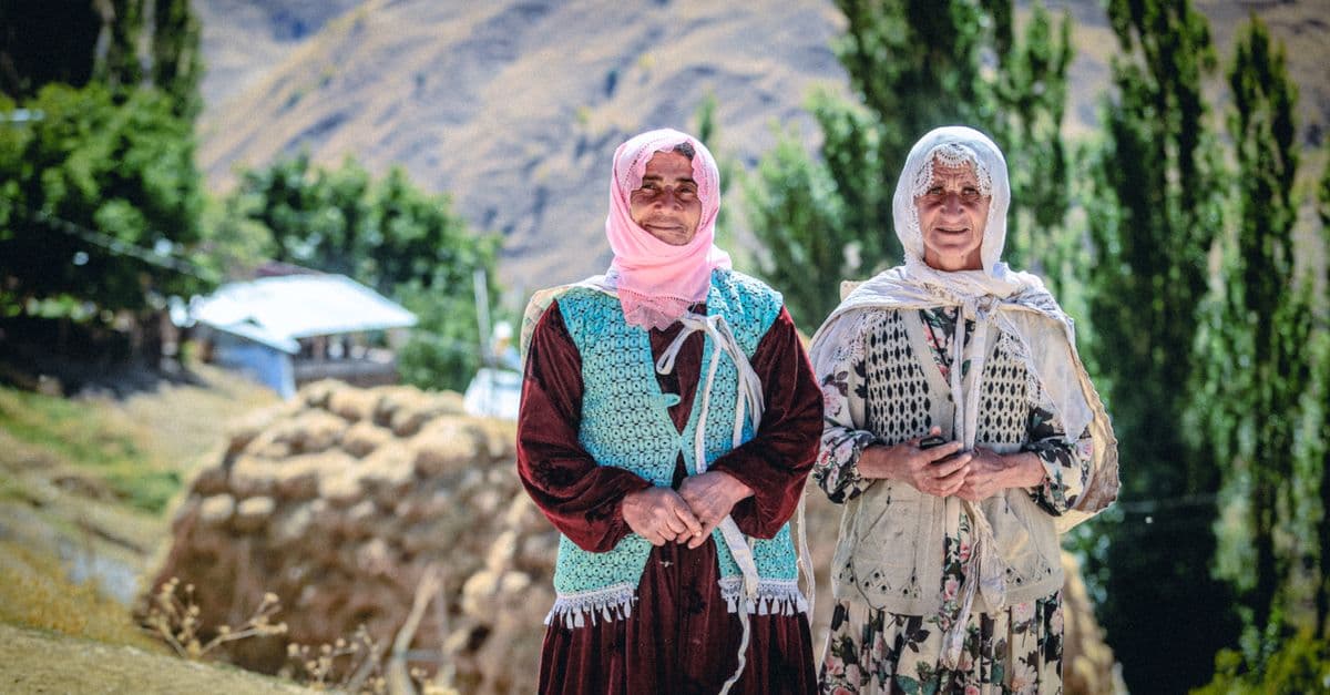 Two elderly women in traditional clothing stand amidst the rural landscape of Van, Türkiye.