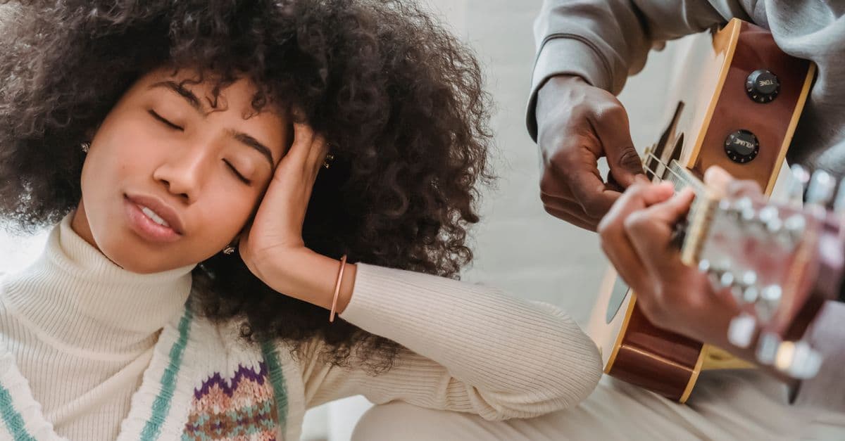 Satisfied African American female with closed eyes listening to anonymous black boyfriend playing song on acoustic guitar while spending time together