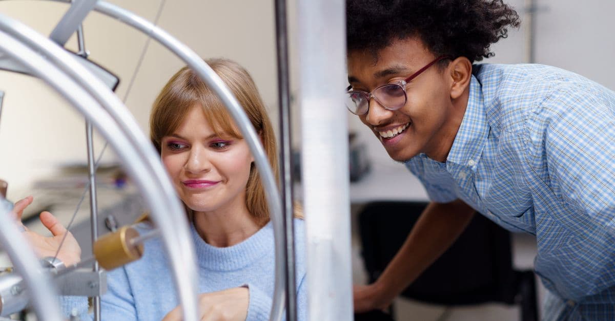 Caucasian woman and African man interacting with physics apparatus in a classroom setting.