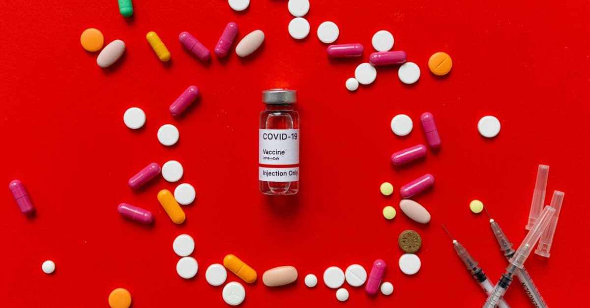 Colorful tablets and COVID-19 vaccine vial on a red background with syringes, symbolizing healthcare.