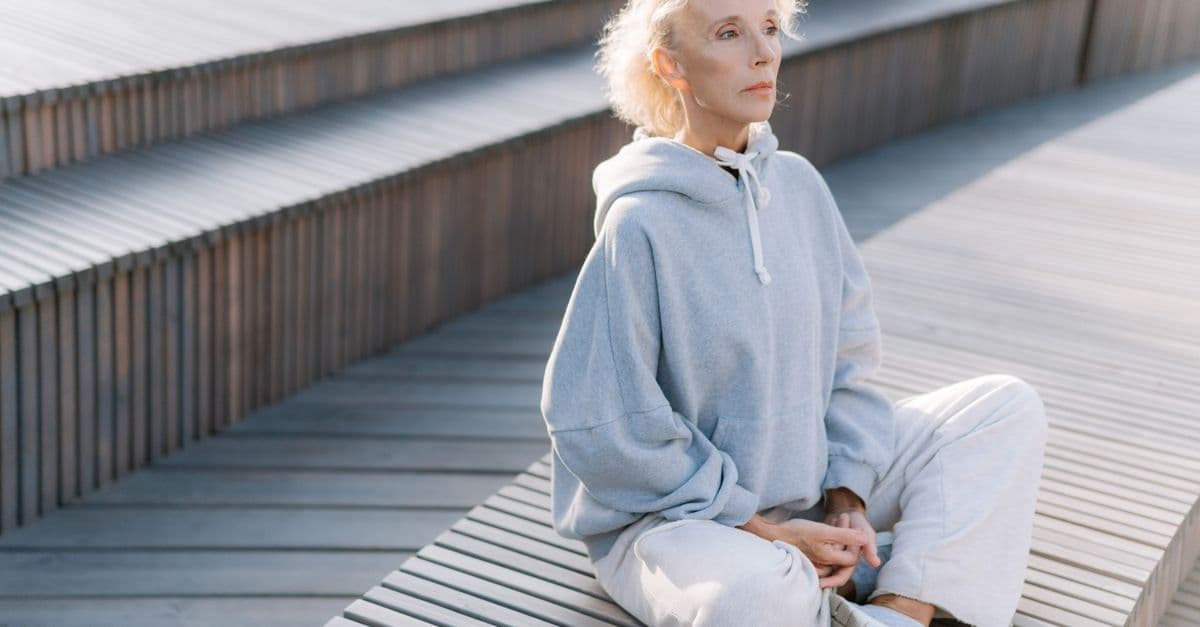 Elderly woman meditating outdoors in sunlight. Relaxation and healthy lifestyle in soft gray sportswear.