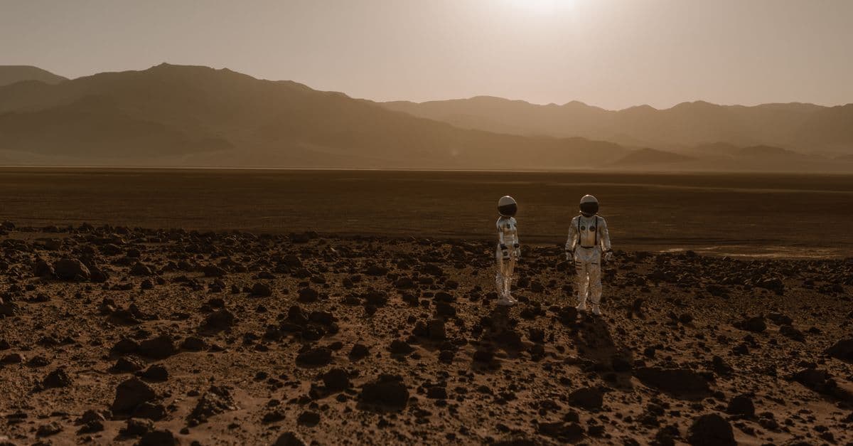 Astronauts in space suits explore a rocky, desert-like terrain resembling Mars at sunset.