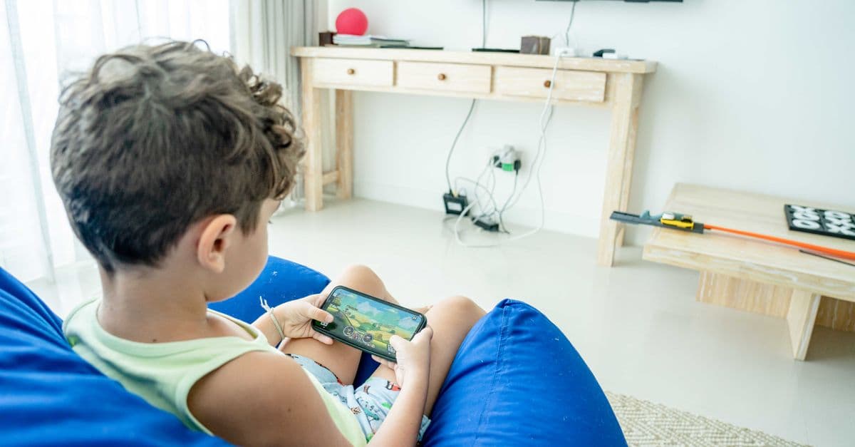 A young boy sitting on a bean bag playing a game on his smartphone in a modern living room.