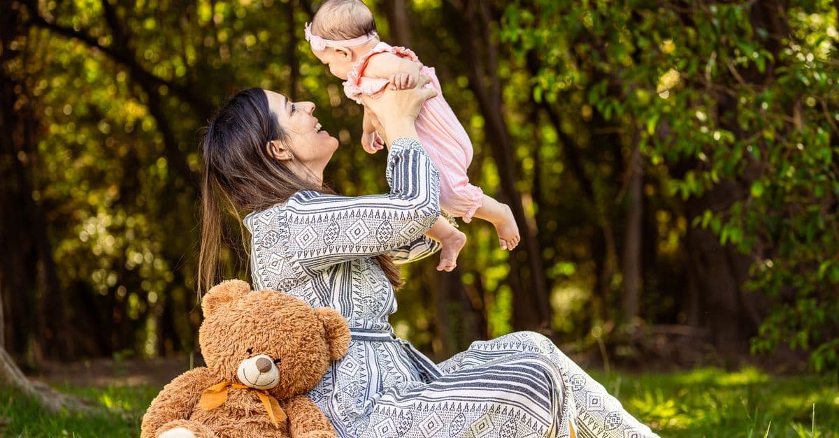 A joyful mother holds her baby up in the forest, surrounded by greenery and sunlight.