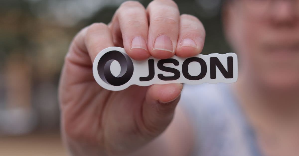 A close-up of a hand holding a JSON logo sticker outdoors, blurred background.