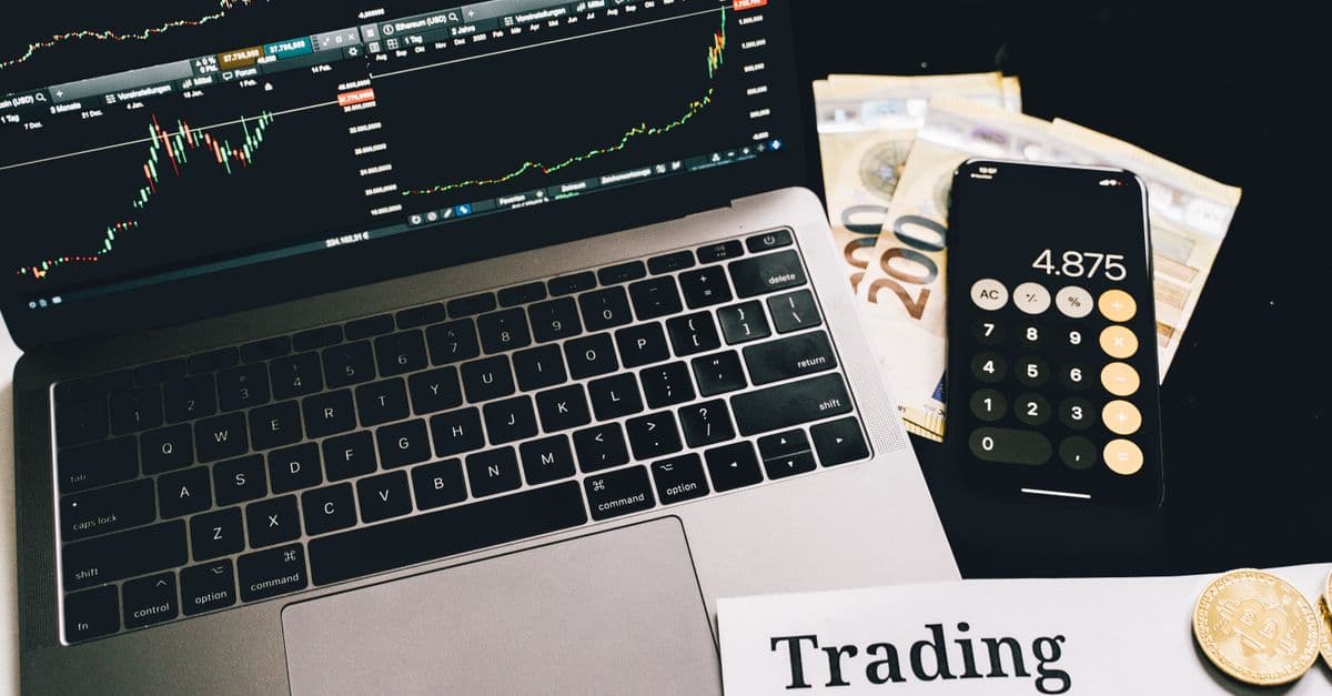 A laptop showing stock market graphs with a smartphone calculator, euro bills, coins, and trading strategies paper.