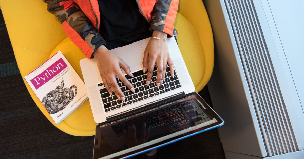 High-angle view of woman coding on a laptop, with a Python book nearby. Ideal for programming and tech content.
