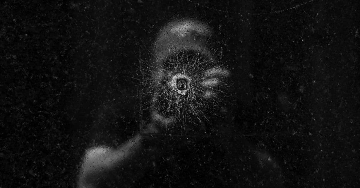 Artistic photograph of a person's reflection seen through a bullet hole on a dark glass surface.