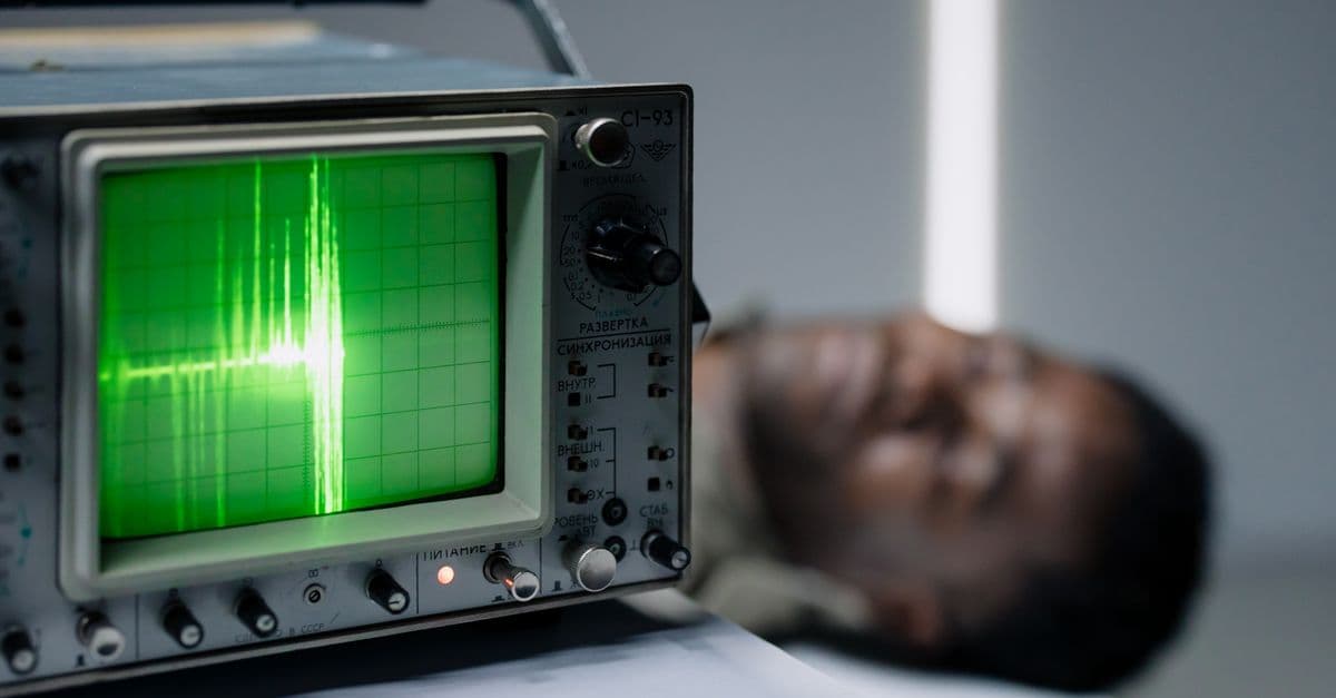 Close-up of a vintage oscilloscope displaying a green waveform next to a blurred person.