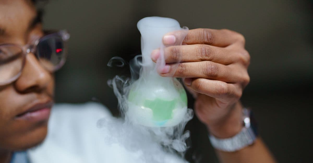 Scientist examining a flask with smoke and green liquid, demonstrating a laboratory chemical reaction.