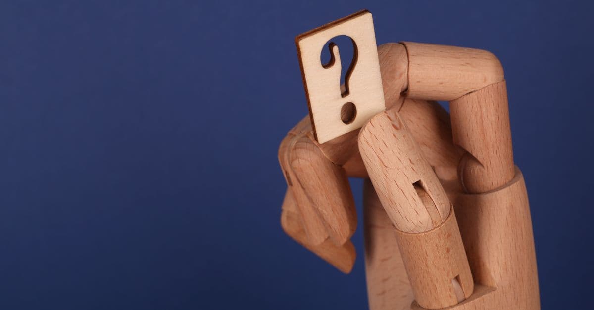 A symbolic wooden hand holding a question mark block against a blue background, representing curiosity and inquiry.