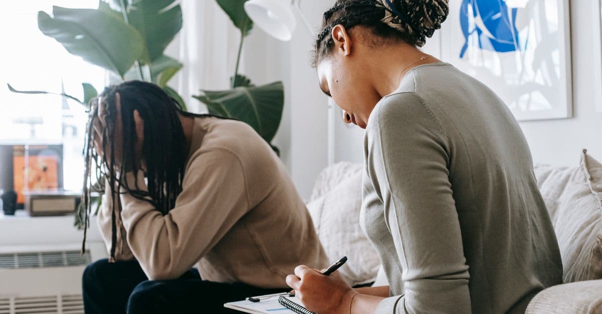Professional female counselor taking notes in notepad while listening to depressed African American male client touching head in dismay in modern psychology center