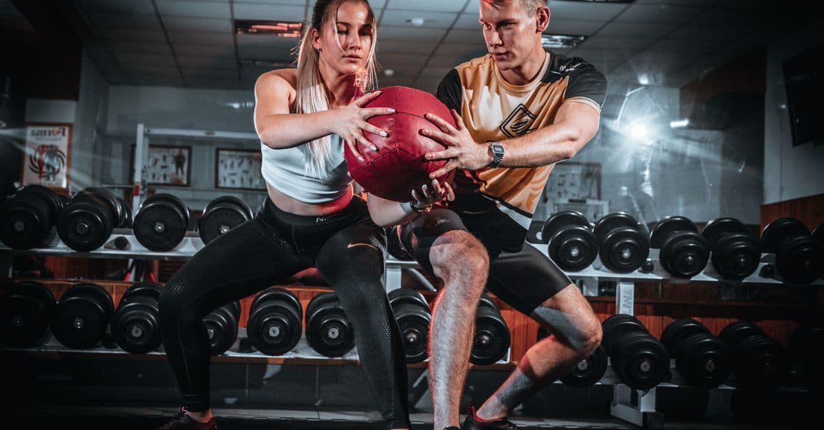 A man and woman perform a tandem workout with a medicine ball in a gym setting.