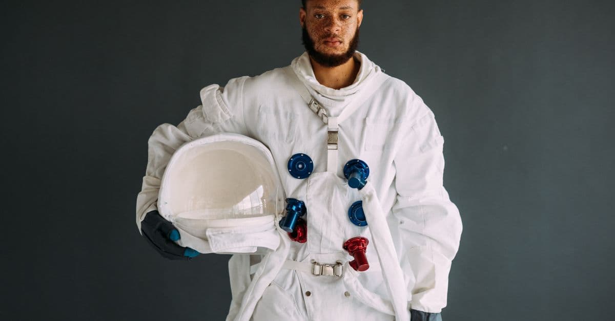A striking portrait of an astronaut in a white space suit holding a helmet on a grey background.
