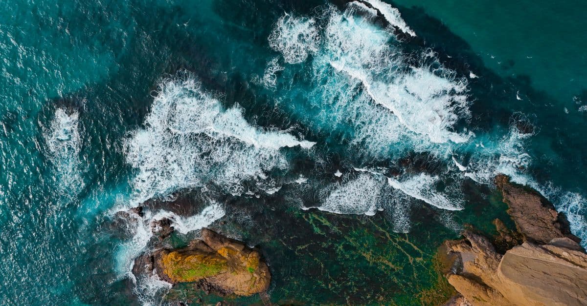 Stunning aerial view of ocean waves crashing onto rocky coastline, showcasing vibrant turquoise waters and rugged cliffs.