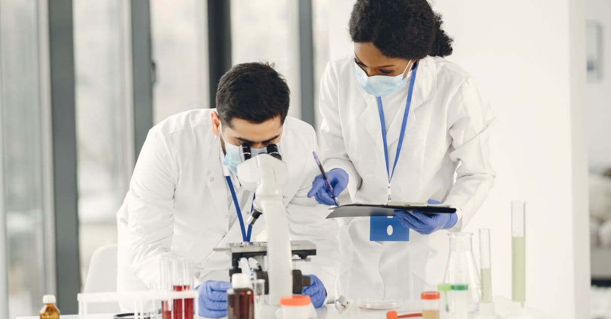 Researchers in lab coats and masks working with microscope and test tubes in a laboratory setting.