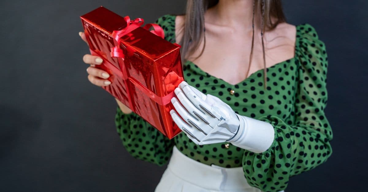 A woman with a prosthetic arm holds a shiny red gift box against a dark background.