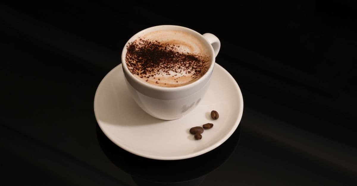 Cup of cappuccino topped with cocoa powder on a saucer with coffee beans.