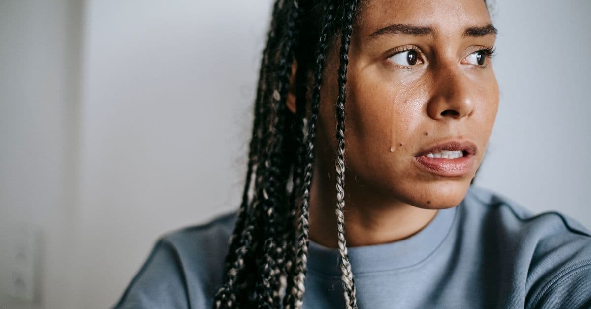 Crop young unhappy African American female with Afro braids in casual clothes looking away in house