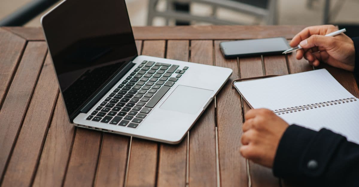 High angle of crop anonymous ethnic male entrepreneur with copybook working at cafeteria table with netbook and smartphone