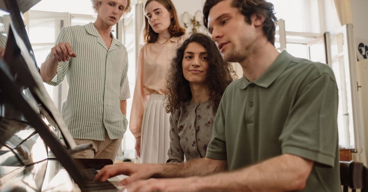 Group of young friends gathered around a piano, playing music indoors.