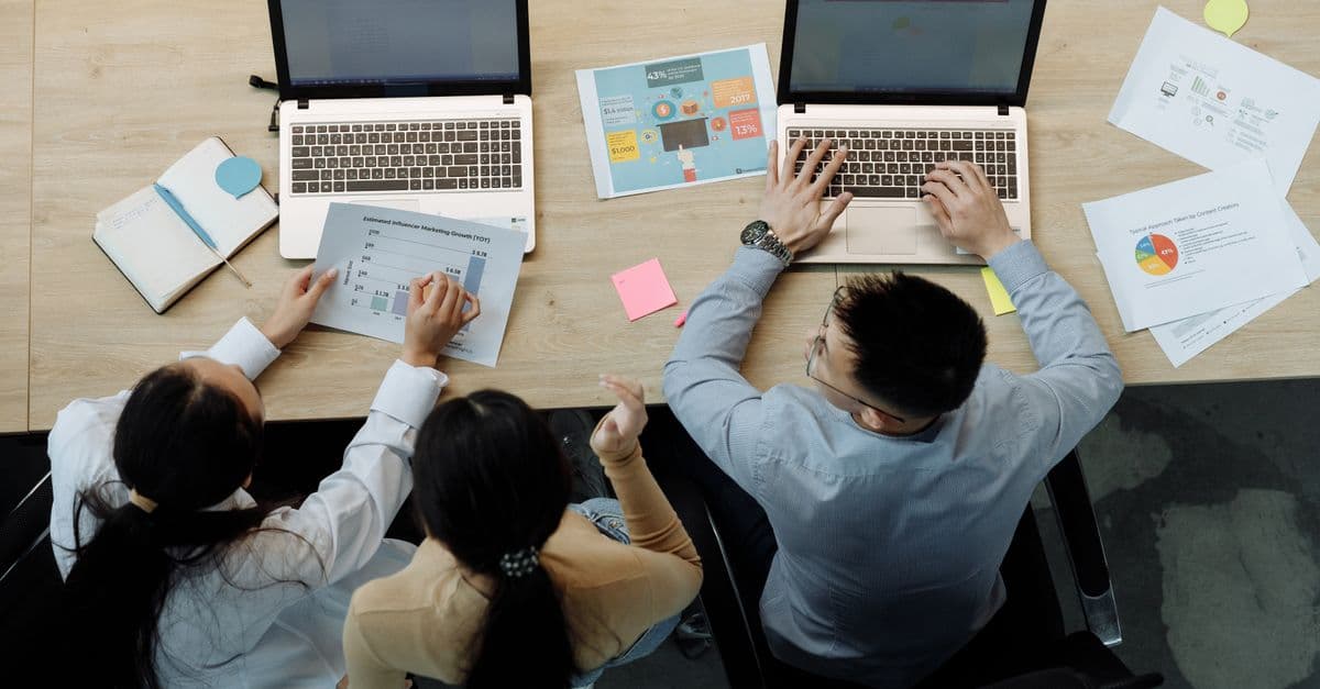 Three colleagues working on laptops and documents at a desk, collaborating on business projects.