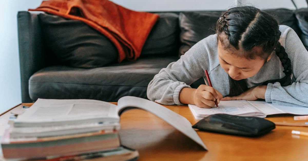 Attentive Asian schoolchild writing in workbook with pen while doing homework at table at home