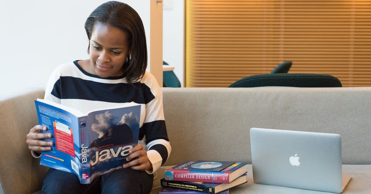 A woman studying Java programming on a sofa with a MacBook and other computer books nearby.